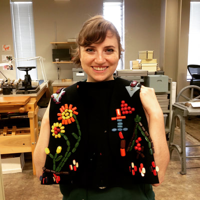 A woman looks at the camera while holding up a dark navy blue felt vest with colored wooden  orange, red, blue, and green beads sewn on in the shape of flowers. 